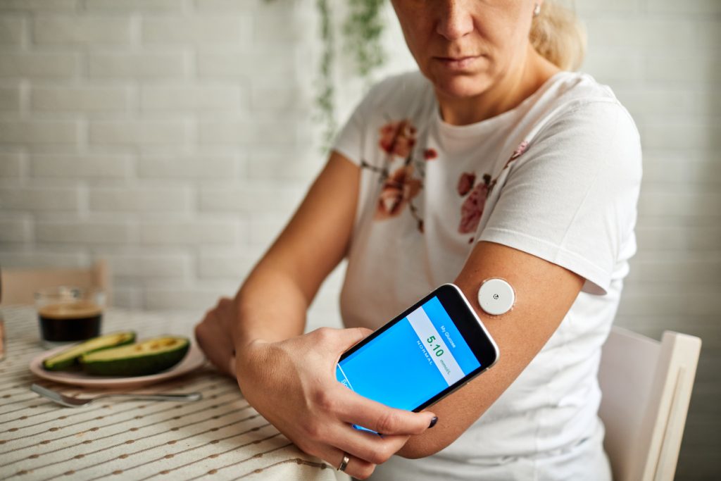 Bild på kvinna i kortärmad vit t-shirt som sitter vid ett matbord och mäter sitt blodsocker med hjälp av sin smartphone och en vit knapp, en sensor som sitter på överarmen
