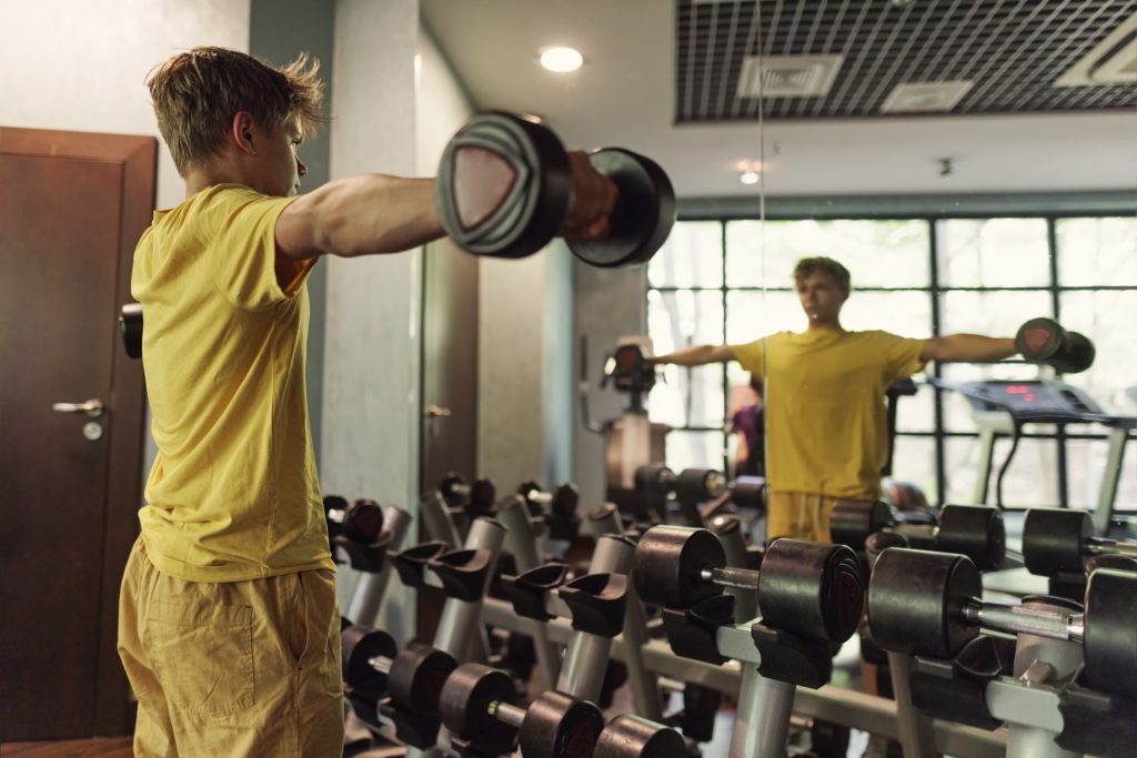 Ung kille lyfter vikter på gym samtidigt som han ser på sig själv i spegel.