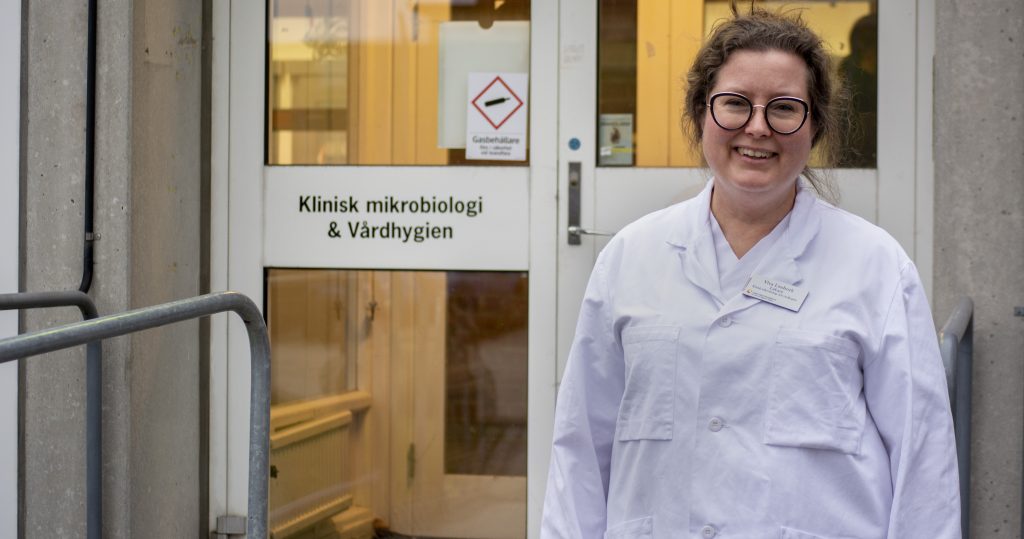 Kvinna i vit labbrock framför glasdörr med skylten "Klinisk mikrobiologi och vårdhygien