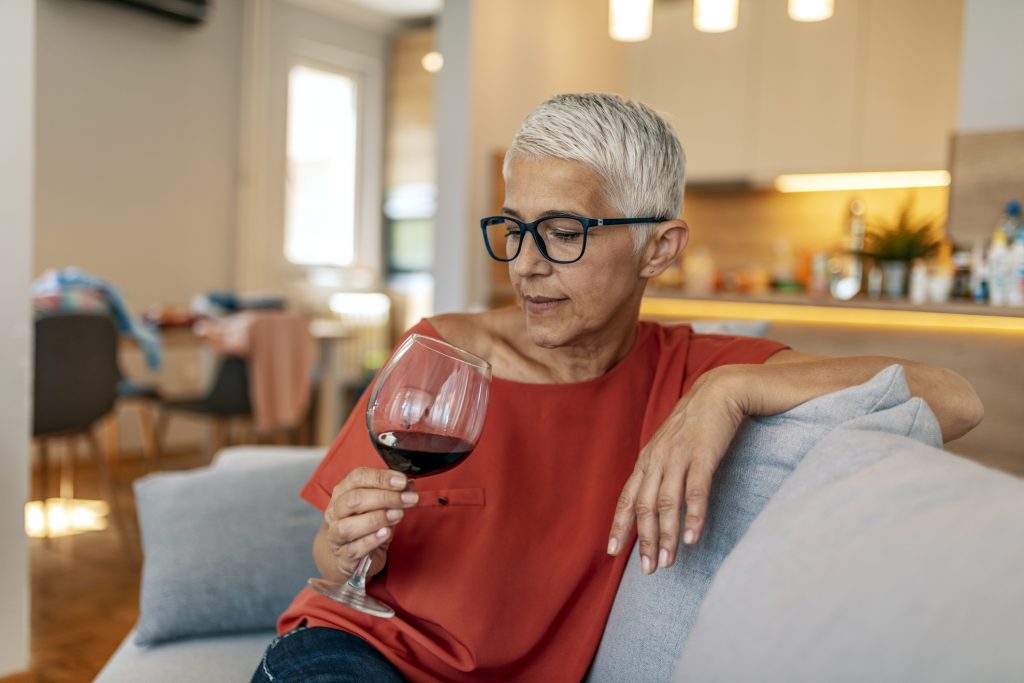 Ensam gråhårig kvinna hemma i soffan med ett glas vin i handen.